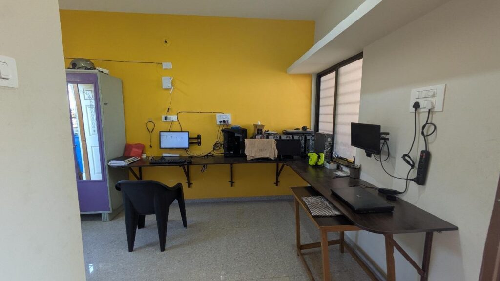 A cluttered home office space featuring multiple computers, monitors, and a long desk. The room has a yellow wall and a purple wardrobe. Good for tech setups, home office ideas, and workspace inspiration. Location: Delhi, India